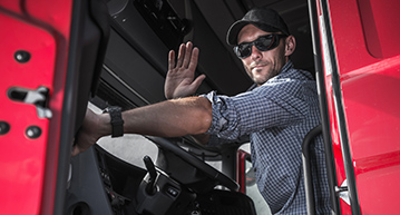 Caucasian male truck driver in a flannel shirt, sunglasses, and dark ball cap sitting in the cab of a semi tractor waving at the camera